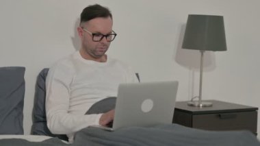 Middle Aged Man Smiling at Camera while using Laptop in Bed