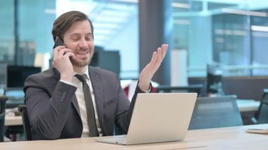 Young Businessman Talking on Phone while using Laptop in Office