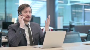 Angry Young Businessman Talking on Phone while using Laptop in Office