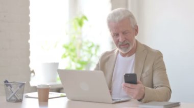 Senior Old Man Browsing Internet on Smartphone while using Laptop in Office 