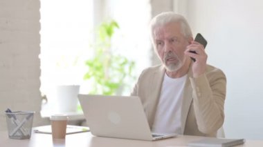 Senior Old Man Talking on Phone while using Laptop in Office
