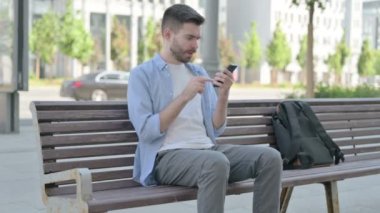 Man Reacting to Loss on Smartphone while Sitting on Bench