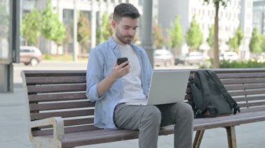 Man Using Smartphone and Laptop while Sitting on Bench