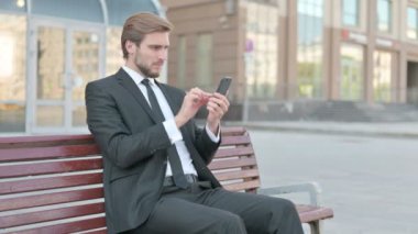 Businessman Reacting to Online Failure via Smartphone while Sitting Outdoor on Bench