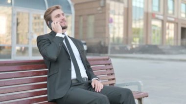 Middle Aged Businessman Talking on Phone while Sitting Outdoor on Bench