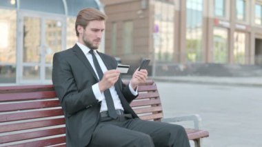 Middle Aged Businessman Reacting to Loss on Smartphone, Sitting Outdoor on Bench