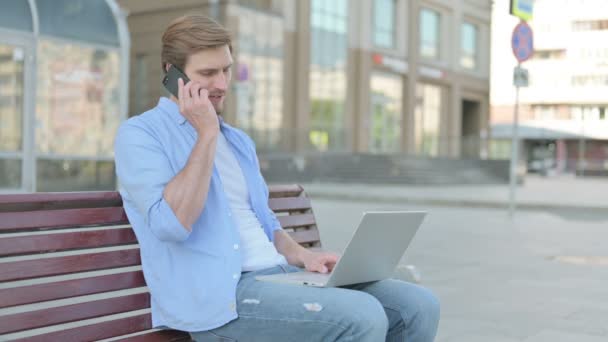 中年男性電話で話しているとベンチに屋外に座っている間にラップトップを使用して — ストック動画