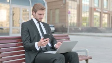 Middle Aged Businessman Using Smartphone and Laptop while Sitting Outdoor on Bench