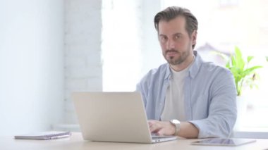 Mature Man Pointing at Camera while using Laptop