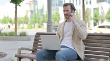 Angry Man with Laptop Talking on Phone while Sitting on Bench