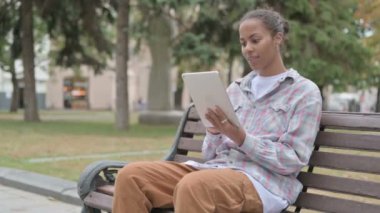 Young African Woman Celebrating Online Win on Tablet while Sitting Outdoor on Bench