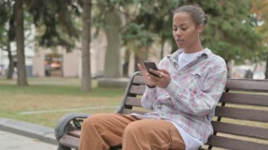 Young African Woman Reacting to Loss on Smartphone while Sitting Outdoor on Bench