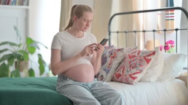 Pregnant Young Woman Using Smartphone while Sitting on Bed