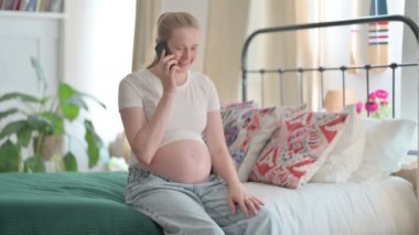 Pregnant Young Woman Talking on Phone while Sitting on Bed