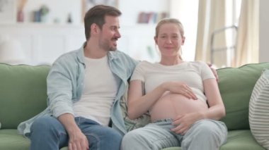 Pregnant Young Woman Talking with Husband while sitting on Sofa