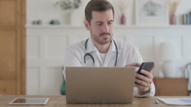 Male Doctor using Smartphone while using Laptop in Clinic 