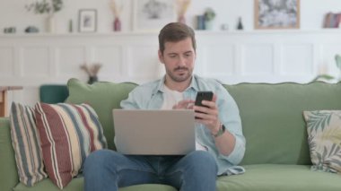 Dizüstü bilgisayarı olan sıradan bir adam koltukta Smartphone kullanıyor. 