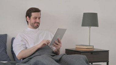 Sakallı genç adam yatakta otururken tablet kullanıyor.