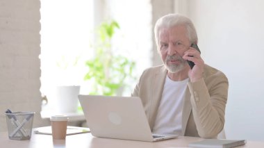 Senior Old Man Talking on Phone while using Laptop in Office