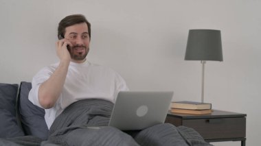 Dizüstü bilgisayarlı Sakallı Genç Adam Yatakta Akıllı Telefonla Konuşuyor