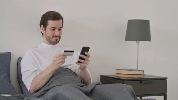 在床上用智能手机在线付款的胡子年轻人 — 图库照片
