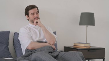 Sakallı genç adam yatakta otururken düşünüyor.