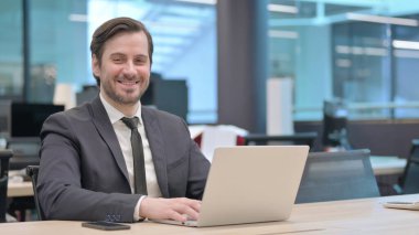 Ofiste dizüstü bilgisayar kullanırken kameraya gülümseyen genç iş adamı