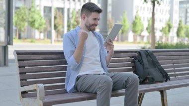 Man Celebrating Online Win on Tablet while Sitting on Bench