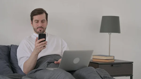 在床上用智能手机的带着手提电脑的胡子年轻人 — 图库照片