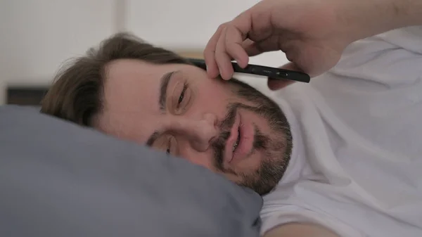 在床上用智能手机说话的胡子年轻人的画像 — 图库照片