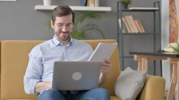 Beard Young Man with Laptop Reading Documents on Sofa