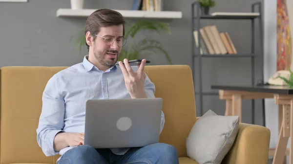 Angry Beard Young Man with Laptop Talking on Smartphone on Sofa