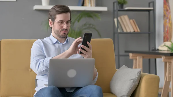 Beard Young Man with Laptop using Smartphone on Sofa