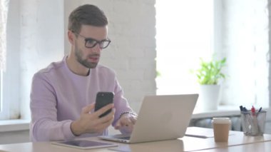 Genç Yetişkin Adam dizüstü bilgisayar kullanırken akıllı telefon kullanıyor 