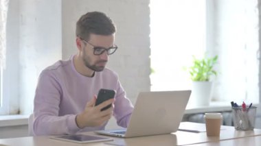 Kızgın Genç Yetişkin Adam Laptop 'ta çalışırken telefonla konuşuyor.