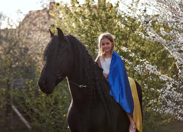 美しい小さなブロンドの女の子乗馬A Friesian馬とともにウクライナの旗 — ストック写真