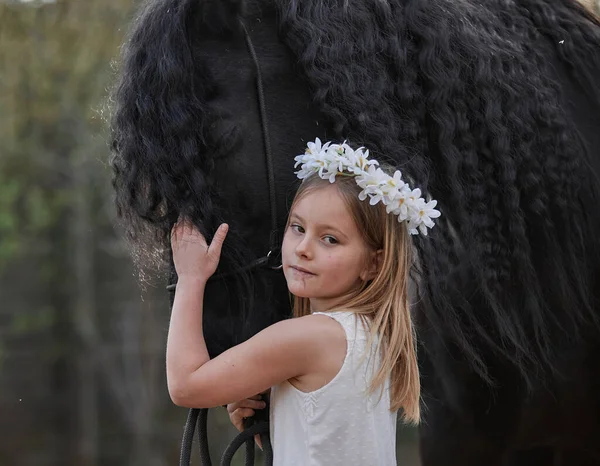 美丽的小女孩 白发苍苍 春天里头戴花环 牵着一匹弗里斯兰马在花园里散步 — 图库照片