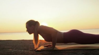 Kumsalda yoga yapan genç sarışın kadın güneşin doğuşunun ya da batışının arka planında