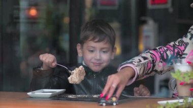 Anne ve oğlu bir kafede oturuyor. Çocuk kek yiyor. Kadın sütlü kahve içiyor.