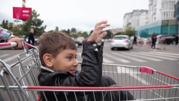 Mama buja syna w wózku sklepowym, niesie go po parkingu w pobliżu supermarketu. — Wideo stockowe