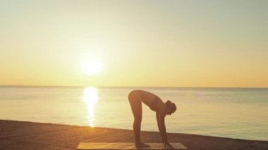 Kumsalda yoga yapan genç sarışın kadın güneşin doğuşunun ya da batışının arka planında