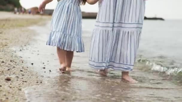 Мать и дочь ходят по пляжу по песку, морская вода мочит их босые ноги. — стоковое видео