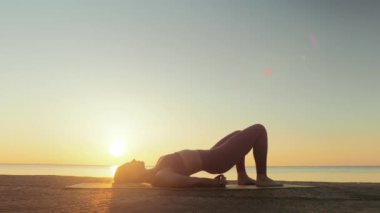 Kumsalda yoga yapan genç sarışın kadın güneşin doğuşunun ya da batışının arka planında
