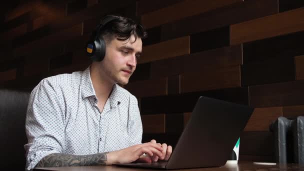 Чоловік брюнетка сидить в кафе за ноутбуком Робочі дослідження, п'є каву Кейси музика в навушниках — стокове відео