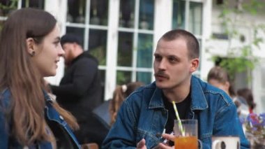 Genç bir çift. Denim giysileri. Eski bir restoranda randevuda, dışarıda. Konuşuyorlar. Sallanıyorlar.
