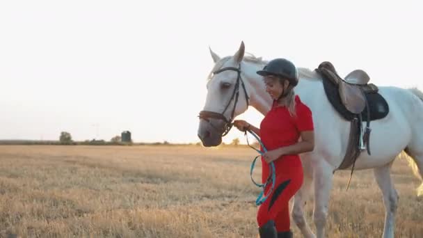 赤い制服を着た女性が白い馬と干し草の畑を歩いている — ストック動画