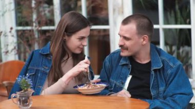 Genç bir çift. Denim giysileri. Eski bir restoranda randevuda, dışarıda. Konuşuyorlar. Sallanıyorlar.