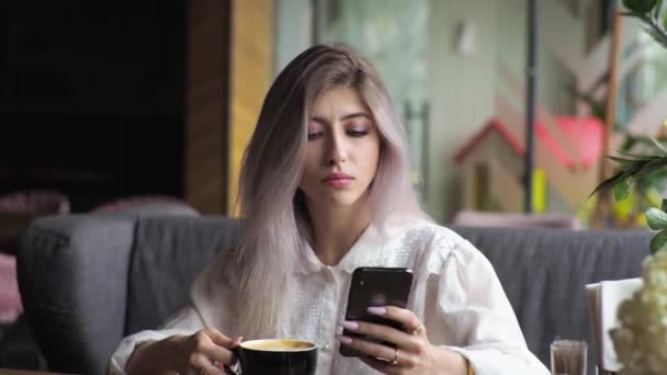 年轻的金发女商人在餐馆的窗户边喝咖啡 看看智能手机吧 在聊天中键入文本消息 — 图库视频影像