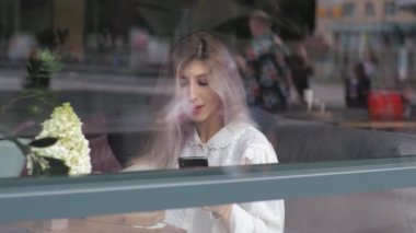 Genç sarışın iş kadını bir restoranın penceresinde kahve içiyor. Akıllı telefona bakıyor. Sohbete bir mesaj yaz. Pencereden bak.