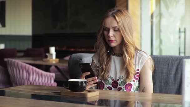 一个穿着休闲装的年轻女子坐在一家咖啡馆的窗边喝咖啡 看着电话 在社交网络上输入文字 — 图库视频影像